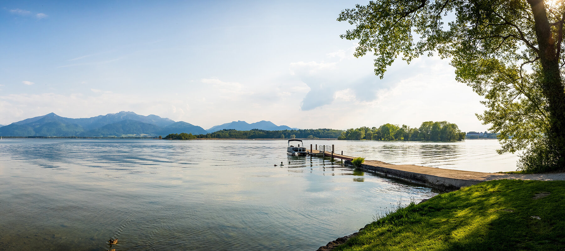 Ein idyllischer See mit ruhigem Wasser und einer kleinen Anlegestelle, an der ein Boot festgemacht ist. Im Hintergrund sind bewaldete Berge und eine baumbestandene Insel zu sehen. Rechts ragen grüne Bäume ins Bild, am Ufer wächst grünes Gras. Der Himmel ist leicht bewölkt und die Szenerie wirkt friedlich und einladend. Ein idyllischer See mit ruhigem Wasser und einer kleinen Anlegestelle, an der ein Boot festgemacht ist. Im Hintergrund sind bewaldete Berge und eine baumbestandene Insel zu sehen. Rechts ragen grüne Bäume ins Bild, am Ufer wächst grünes Gras. Der Himmel ist leicht bewölkt und die Szenerie wirkt friedlich und einladend.