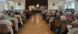 Ein voll besetzter Saal mit älteren Menschen, die auf Stühlen sitzen und einer Musikaufführung auf einer Bühne zuhören. Auf der Bühne befinden sich mehrere Musiker und ein Dirigent. Die Perspektive ist von hinten, sodass das Publikum im Vordergrund und die Bühne im Hintergrund zu sehen sind.