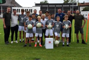 Eine Fußballmannschaft bestehend aus mehreren Jungen, einem Mädchen und mehreren Erwachsenen posiert mit Fußbällen und in grauen Trikots auf einem Fußballplatz, im Hintergrund Gebäude. Copyright: Franz Ruprecht.