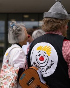 Mehrere Personen im Freien, im Fokus steht eine Person mit einem schwarzen Kleidungsstück, auf dem das Logo der „Klinikclowns“ mit einem stilisierten lachenden Gesicht und roter Nase zu sehen ist. Diese Person trägt außerdem eine Gitarre auf dem Rücken. Im Hintergrund sind weitere Personen unscharf erkennbar.