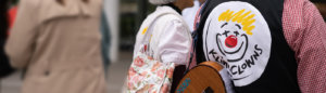 Mehrere Personen im Freien, im Fokus steht eine Person mit einem schwarzen Kleidungsstück, auf dem das Logo der „Klinikclowns“ mit einem stilisierten lachenden Gesicht und roter Nase zu sehen ist. Diese Person trägt außerdem eine Gitarre auf dem Rücken. Im Hintergrund sind weitere Personen unscharf erkennbar.