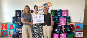 Drei Frauen stehen nebeneinander und halten gemeinsam ein großes Spendenschild, auf dem eine Spende von 10.000 Euro der Roland und Renate Gruber Stiftung an die Aktion Aufwind verkündet wird. Im Hintergrund sind zahlreiche bunte Schulranzen vor einer Wand mit Aufwind-Logos gestapelt. Alle drei Frauen lächeln in die Kamera.