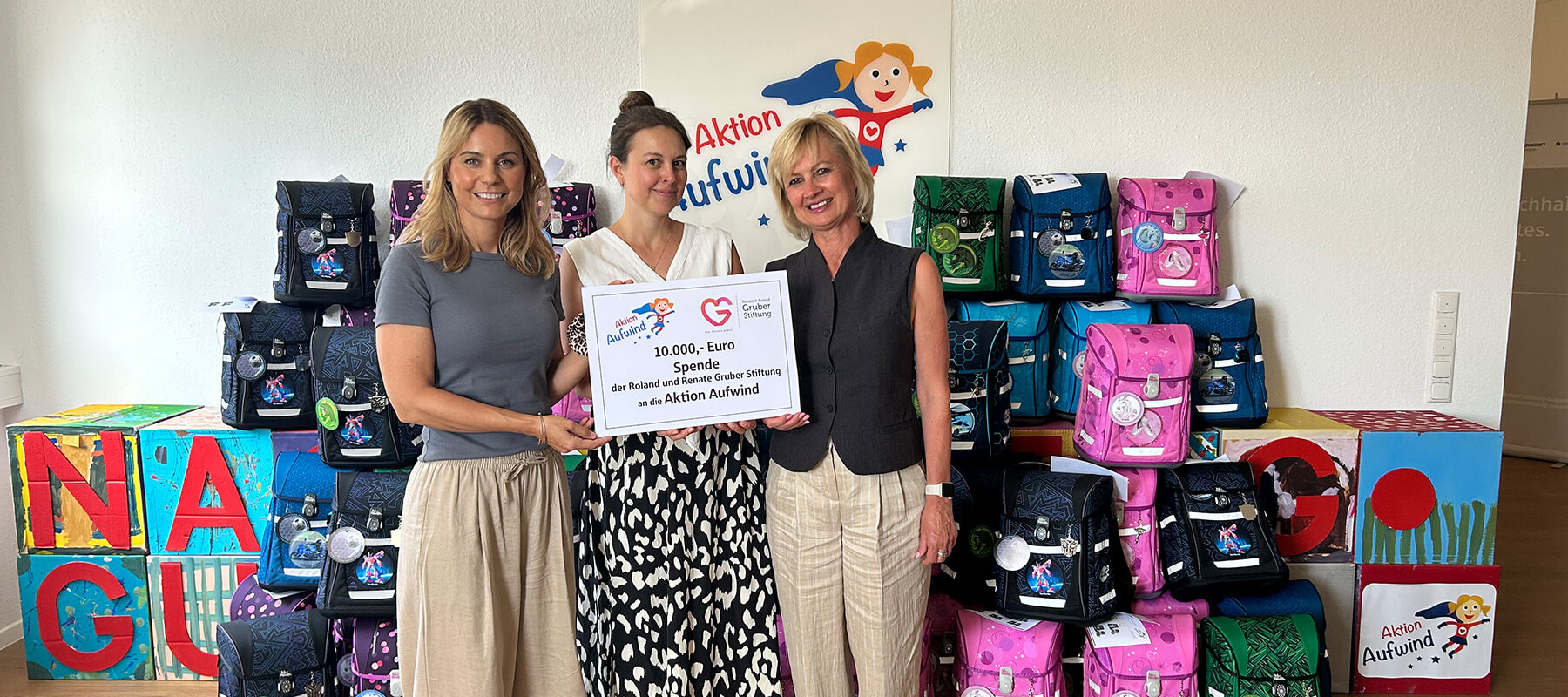 Drei Frauen stehen nebeneinander und halten gemeinsam ein großes Spendenschild, auf dem eine Spende von 10.000 Euro der Roland und Renate Gruber Stiftung an die Aktion Aufwind verkündet wird. Im Hintergrund sind zahlreiche bunte Schulranzen vor einer Wand mit Aufwind-Logos gestapelt. Alle drei Frauen lächeln in die Kamera.