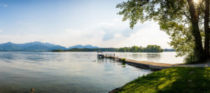 Ein idyllischer See mit ruhigem Wasser und einer kleinen Anlegestelle, an der ein Boot festgemacht ist. Im Hintergrund sind bewaldete Berge und eine baumbestandene Insel zu sehen. Rechts ragen grüne Bäume ins Bild, am Ufer wächst grünes Gras. Der Himmel ist leicht bewölkt und die Szenerie wirkt friedlich und einladend. Ein idyllischer See mit ruhigem Wasser und einer kleinen Anlegestelle, an der ein Boot festgemacht ist. Im Hintergrund sind bewaldete Berge und eine baumbestandene Insel zu sehen. Rechts ragen grüne Bäume ins Bild, am Ufer wächst grünes Gras. Der Himmel ist leicht bewölkt und die Szenerie wirkt friedlich und einladend.
